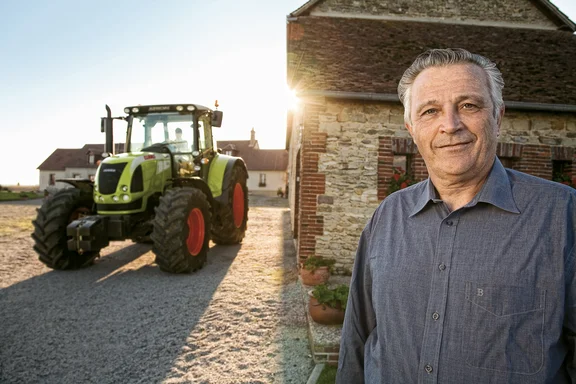 Mann rechts vor einem Haus, links steht ein grüner Trecker